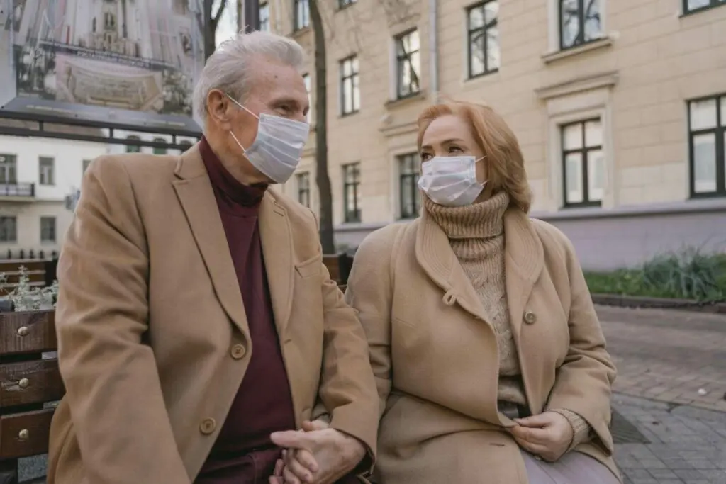 Elderly couple sits on street bench holding hands in camel overcoats and masks.